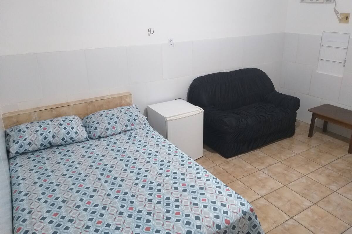 Quarto pequeno e pouco mobiliado, com uma cama, uma cadeira, uma mesinha e um armário branco.