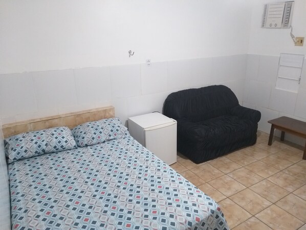 A small, sparsely furnished room with a bed, a chair, a small table, and a white cabinet.