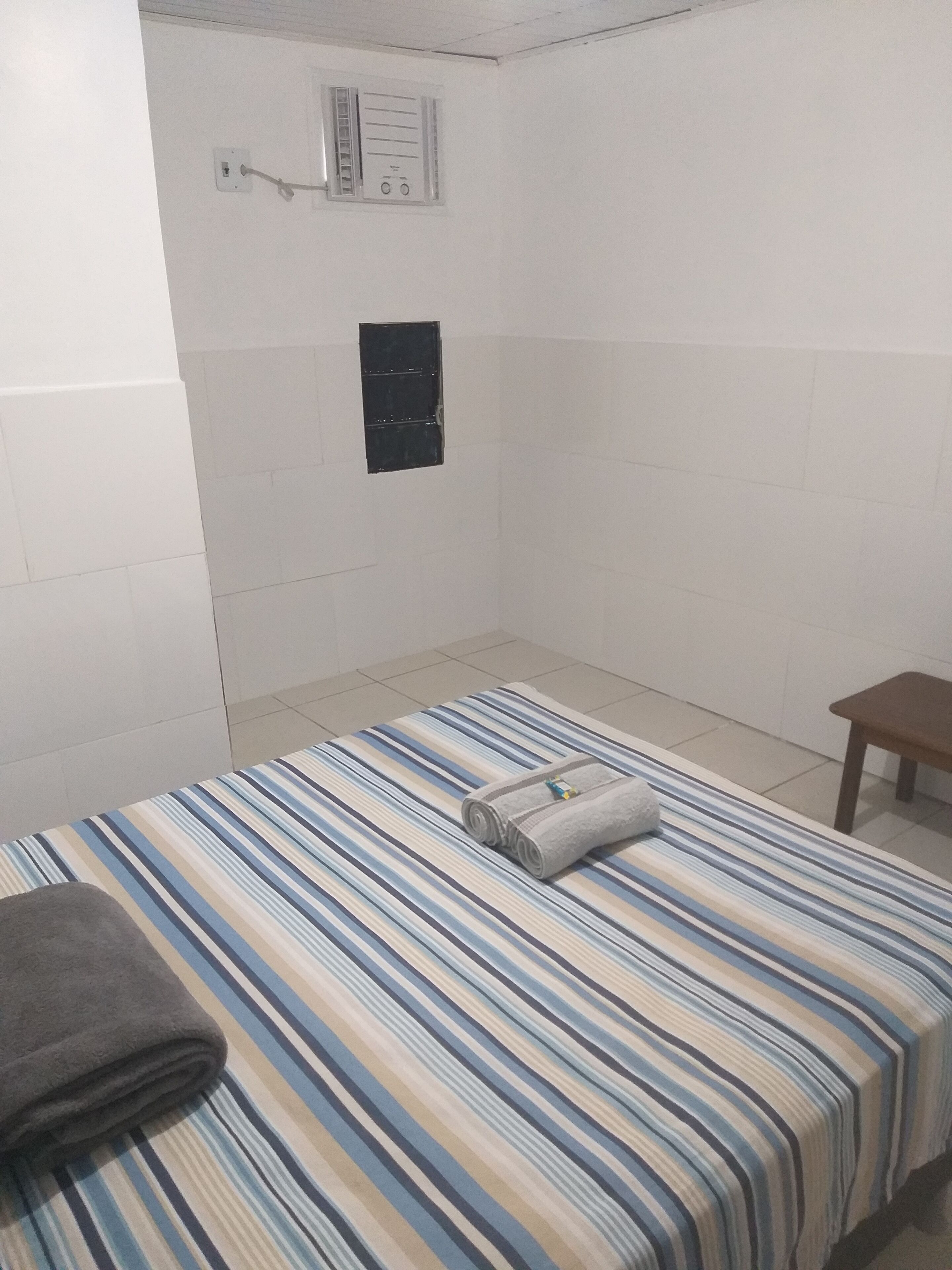 A room with a striped bedspread, a small wooden bench, and a wall-mounted control panel.