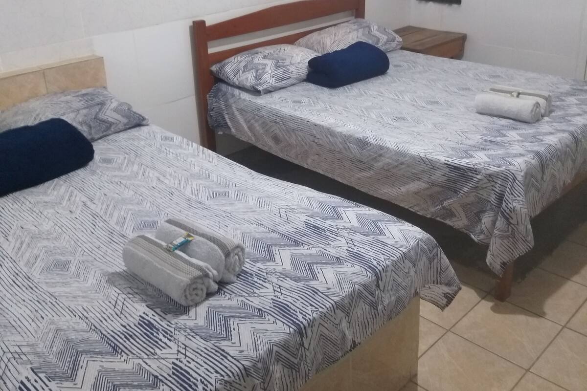 Quarto com duas camas, cada uma com roupa de cama e almofadas estampadas. Há uma pequena mesa de cabeceira de madeira entre as camas, e um aquecedor fixado na parede acima de uma delas.