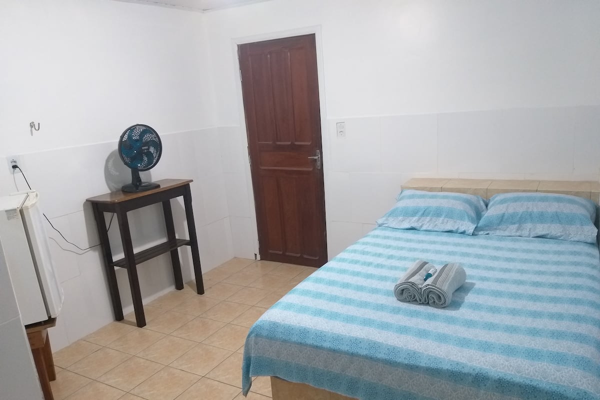 Quarto com cama, porta de madeira e uma pequena mesa com um ventilador.