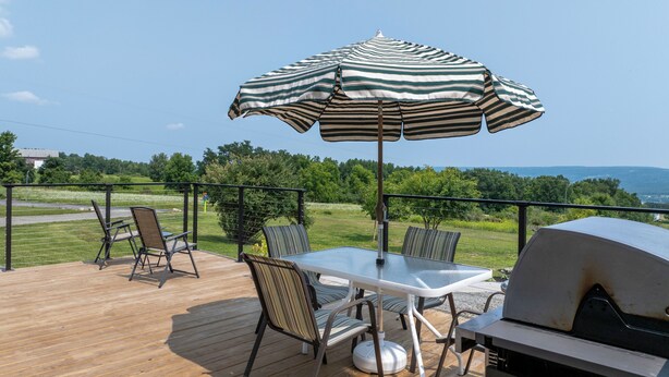 A wooden deck with a table and chairs under a striped umbrella, a grill, and a view of a green landscape. - Lake & Vineyard View (Pulteney)