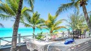 A wooden deck with a hammock and a table, overlooking a beach with palm trees and turquoise water. - Zen Exuma: Stunning Beachfront Villa w/ NEW Rooftop Lounge, Spa, Cabana & Pool! (Moss Town)