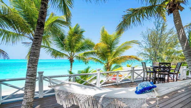 Une terrasse en bois avec un hamac et une table, donnant sur une plage bordée de palmiers et baignée d’une eau turquoise.