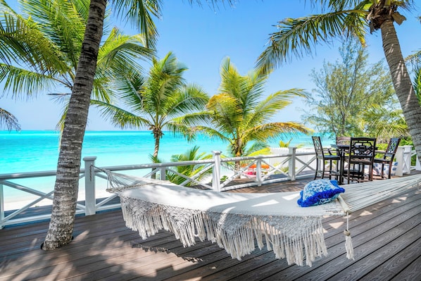 Une terrasse en bois avec un hamac et une table, donnant sur une plage bordée de palmiers et baignée d’une eau turquoise.