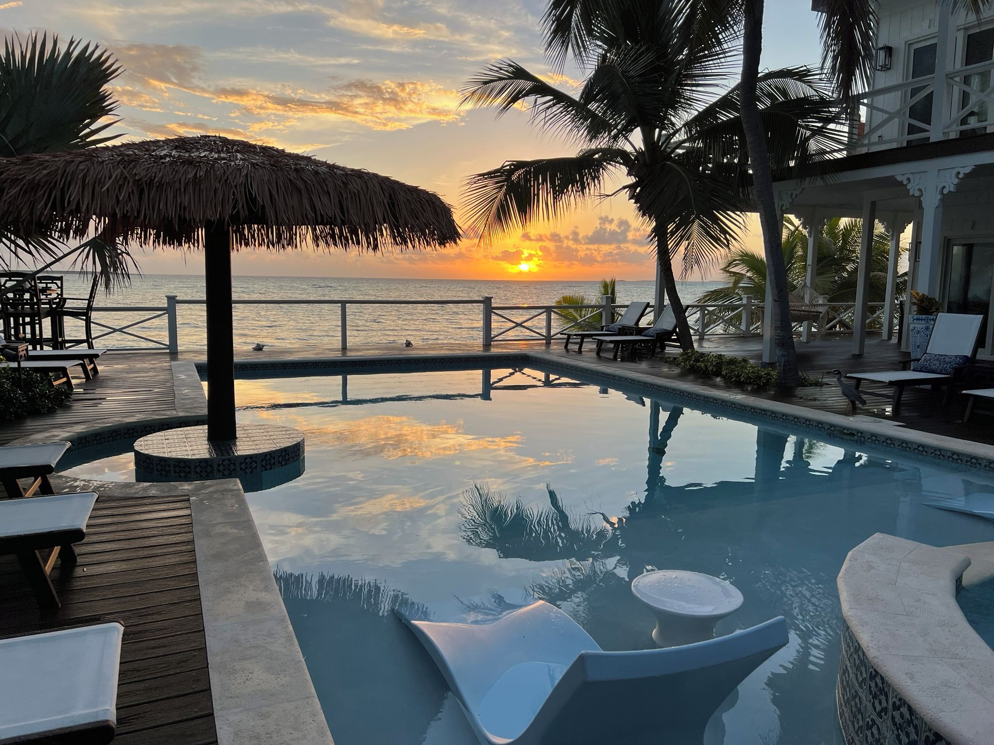 A pool with lounge chairs and a thatched umbrella, overlooking the sea at sunset.