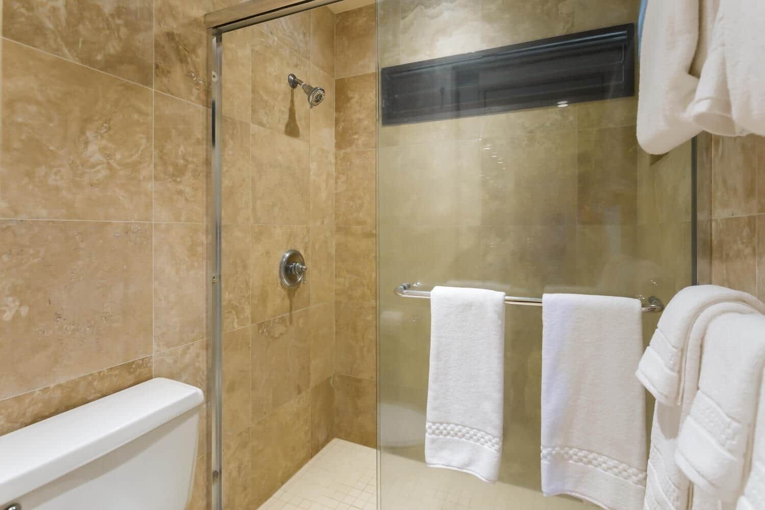 Ducha envuelta en vidrio con cabezal de ducha de mano, puerta de cristal y un perchero con dos toallas blancas.