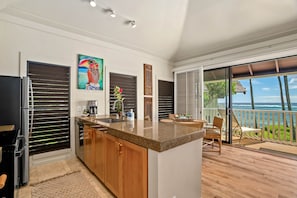 A modern kitchen with a large island, wooden cabinets, and a view to a balcony with outdoor seating. - KP9 Beachfront Ocean View | AC • Pool • Modern (Koloa)