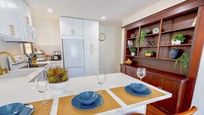 Una cocina moderna con armarios blancos, una mesa de comedor de madera con platos azules, y un estante decorativo con plantas y utensilios de cocina.
