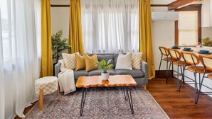 Una sala de estar con sofá gris, cortinas amarillas, una pequeña mesa con una planta, y un comedor con sillas y mesa.