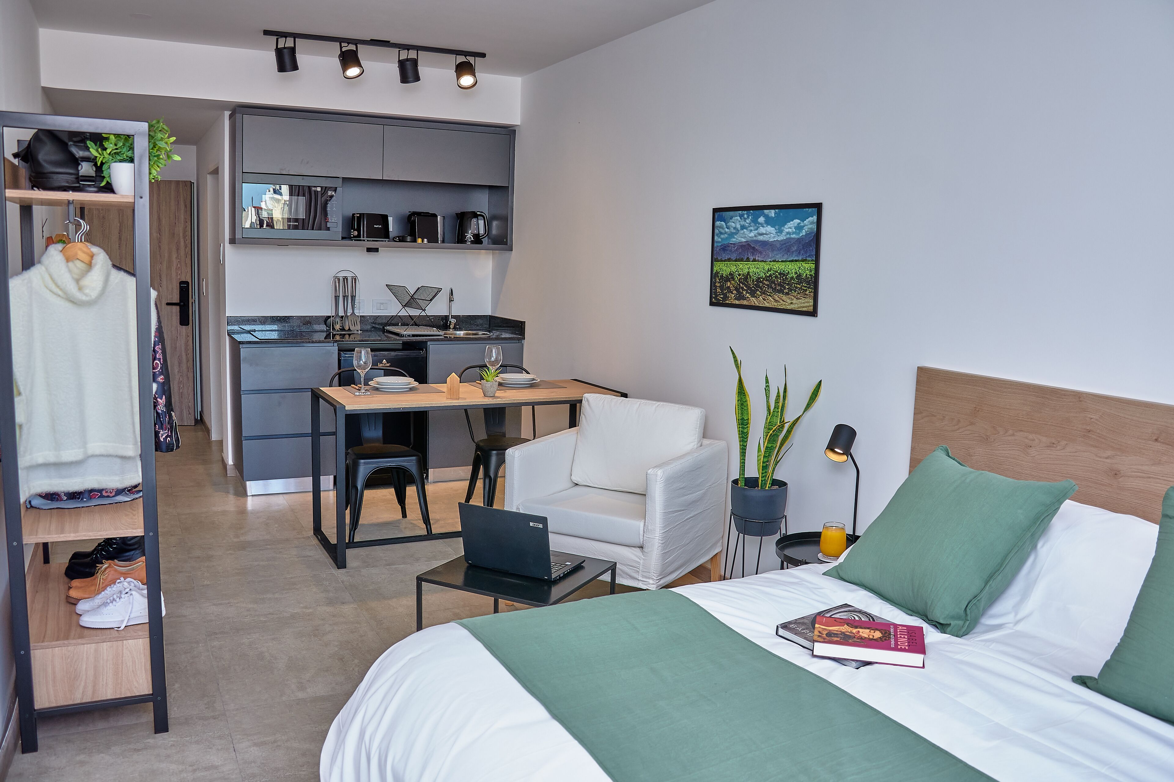 Una habitación de hotel moderna con una cama, un escritorio, una silla, una computadora portátil, una estantería y una pequeña cocina.