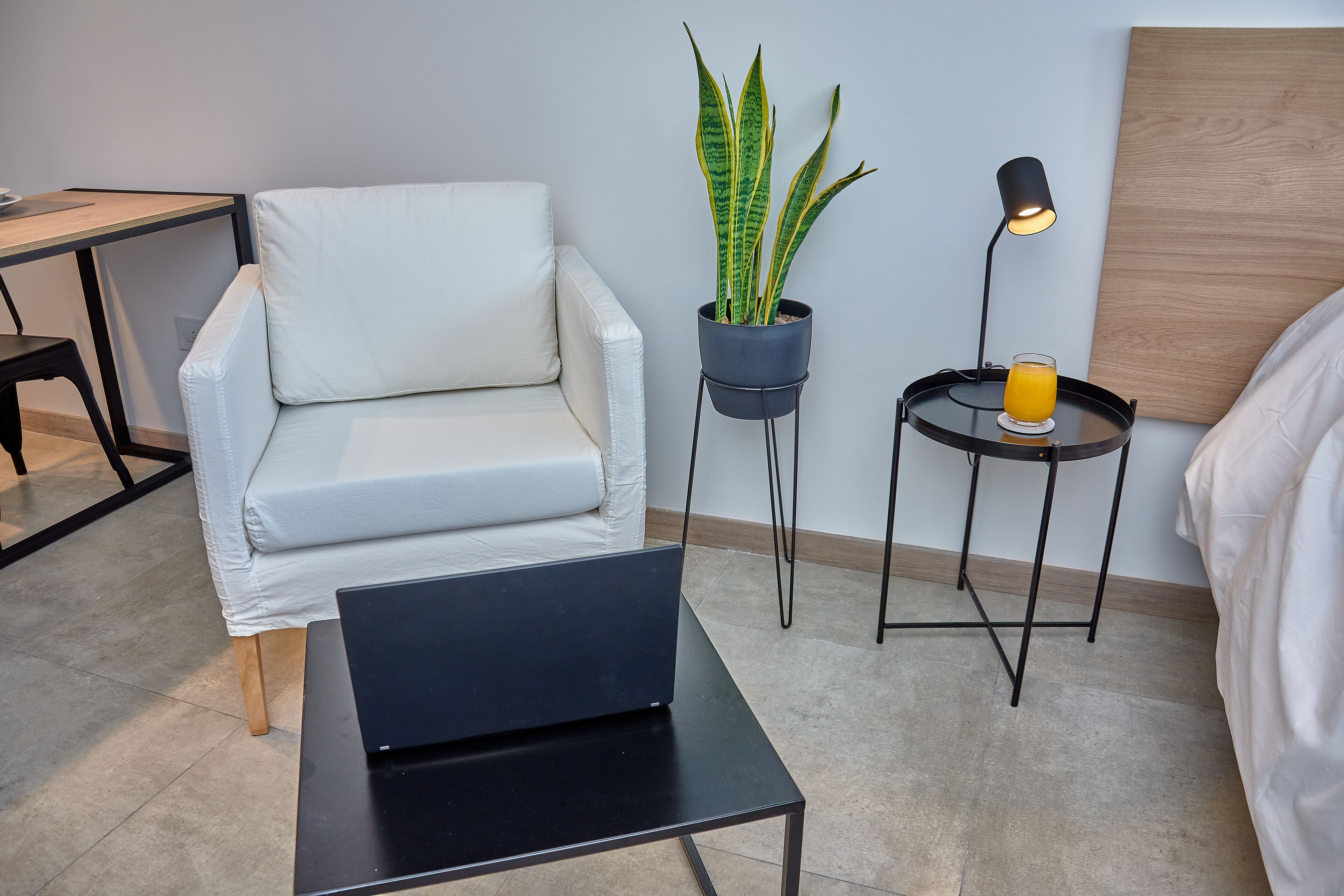 Une chambre moderne avec un canapé blanc, une petite table noire, une plante en pot, une table d’appoint avec une lampe et un verre de jus d’orange, et un lit avec une literie blanche.