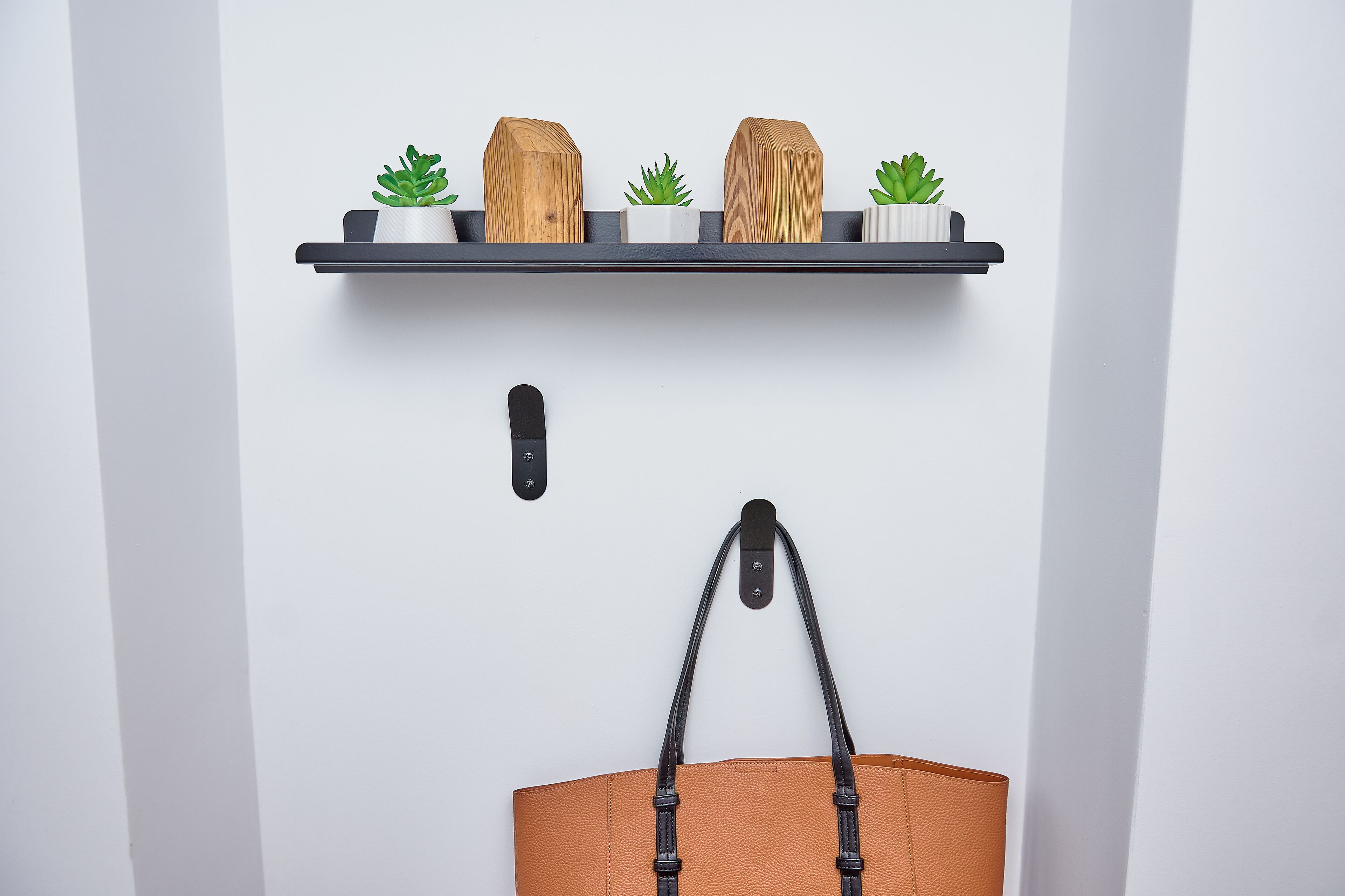 Una puerta blanca con un picaporte negro, un estante negro encima que sostiene tres bloques de madera y dos pequeñas macetas, y una bolsa marrón colgada de un gancho debajo.