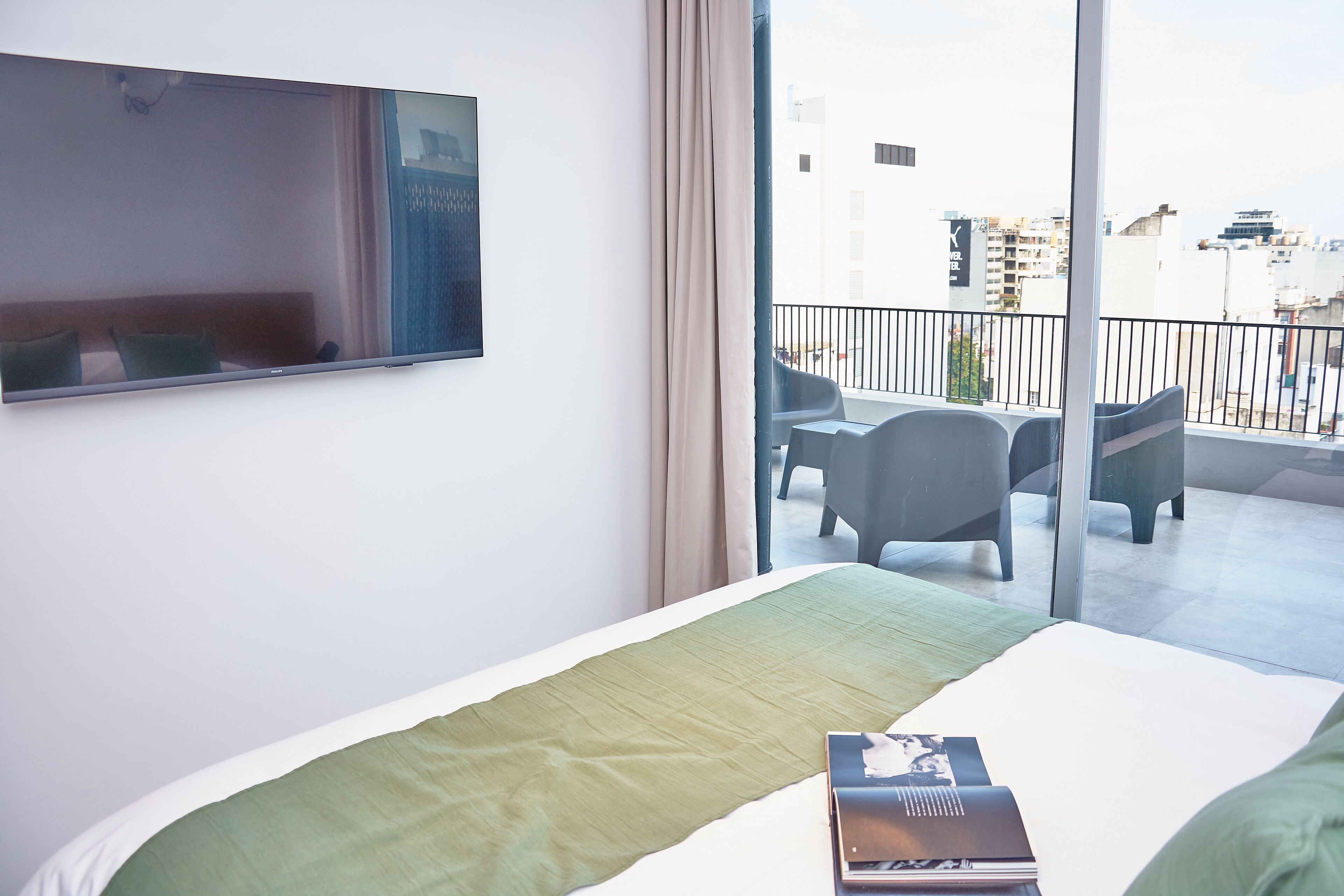 A hotel room with a bed, a flat-screen TV, a balcony with seating, and a view of the city.
