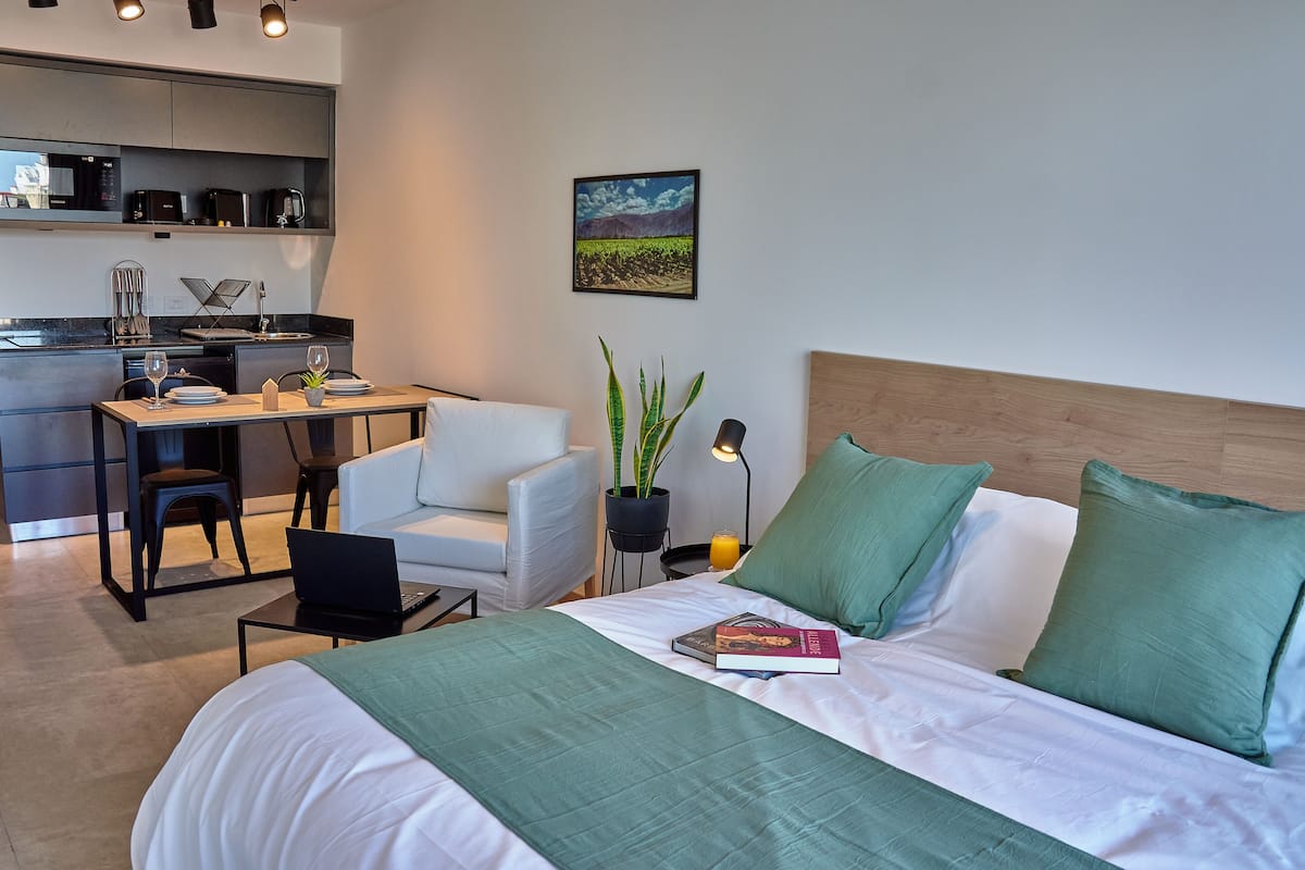 A modern hotel room with a bed, a desk, a chair, a potted plant, and a painting on the wall.