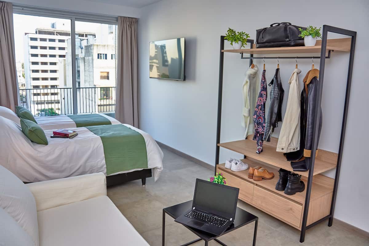 A modern hotel room with a bed, a desk with a laptop, a clothing rack, and a view of the city through the window.