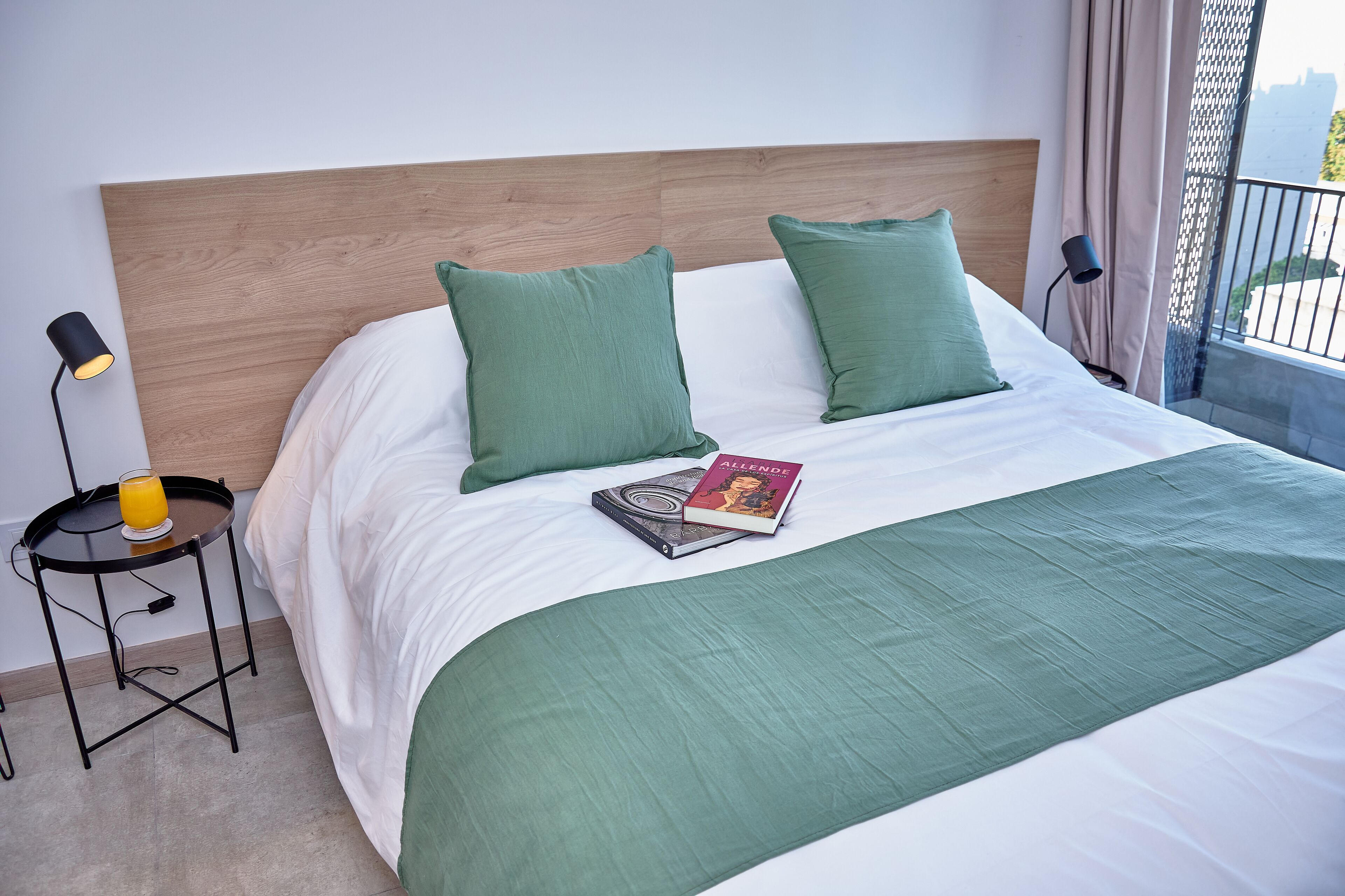 A neatly made bed with green pillows, a book, and a glass of orange juice on a bedside table.