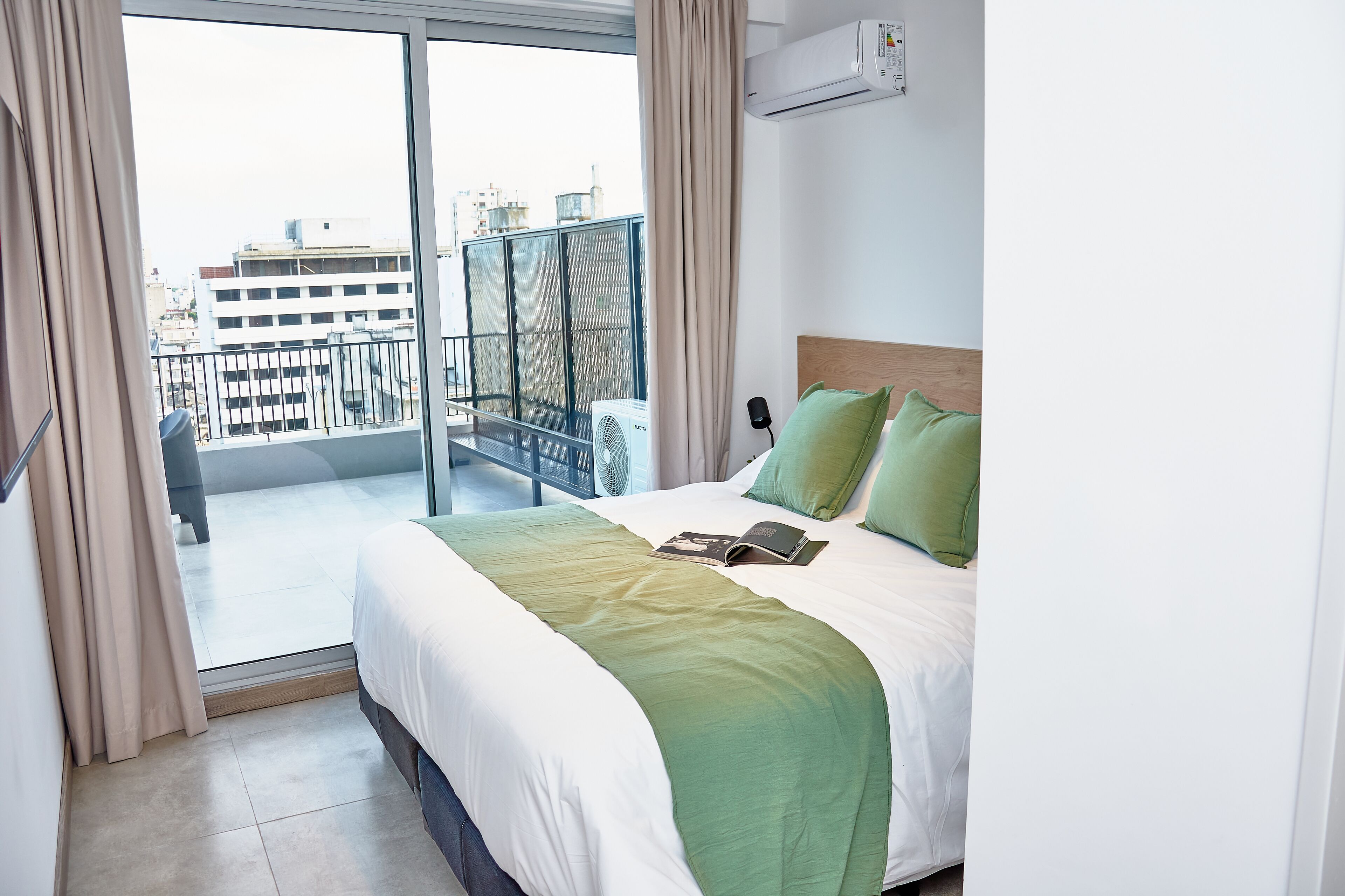 A hotel room with a large bed, a view of the city through a balcony, and an air conditioning unit on the wall.