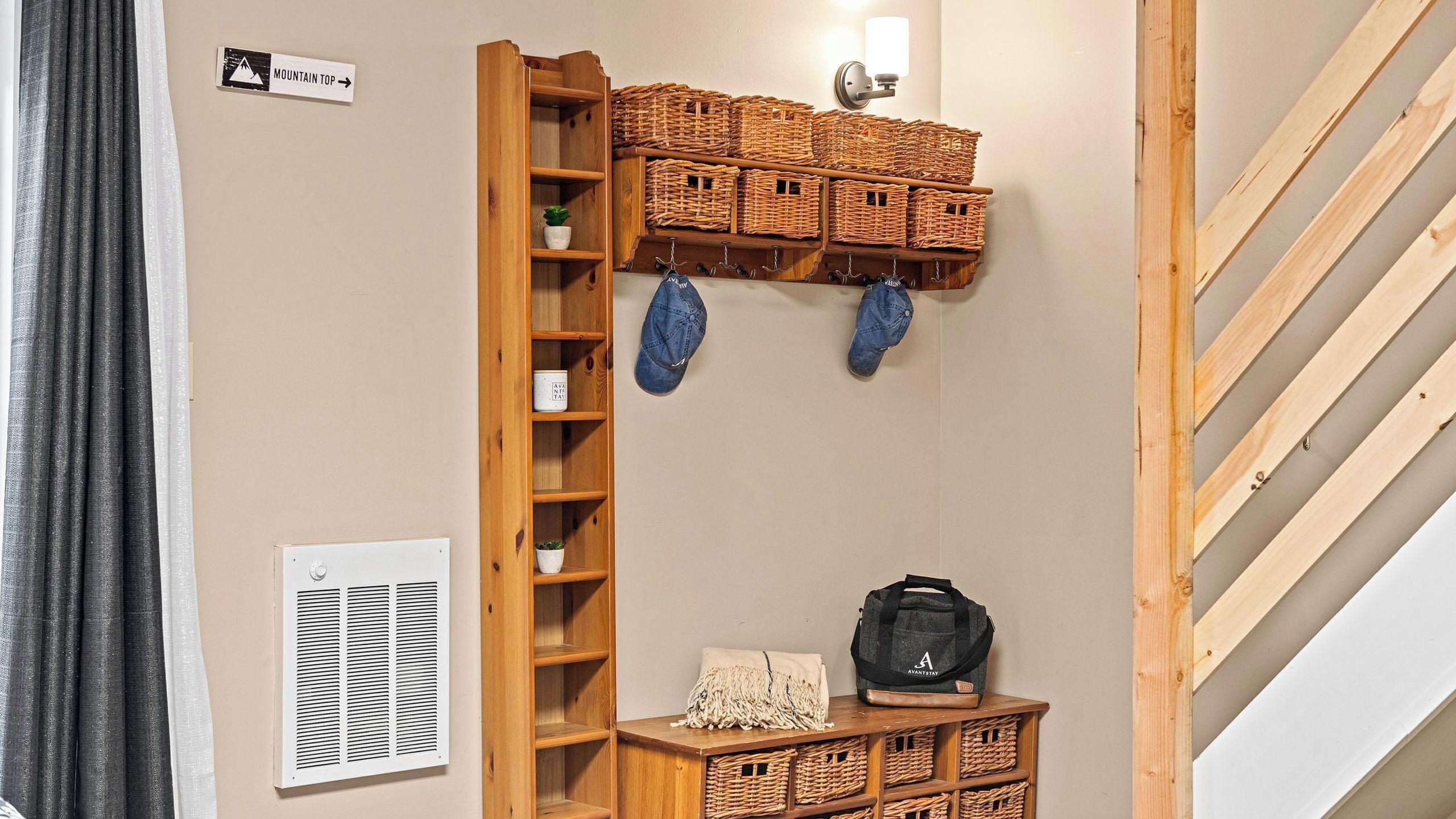 Una habitación con estante de madera con cestas, luz fijada en la pared, perchero con sombreros, una mesita con un bolso y un letrero que dice 'MOUNTAIN TOP'.