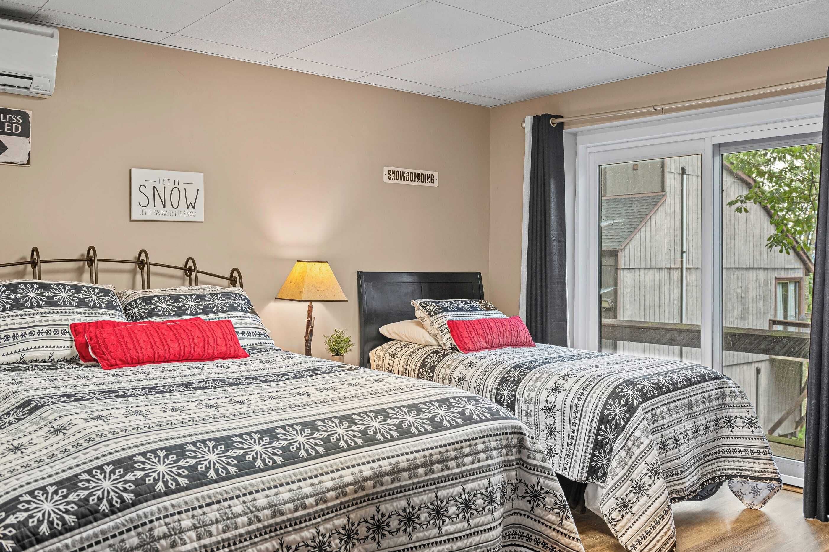 A hotel room with two beds, each with patterned bedding and red pillows. There is a wall-mounted air conditioner and a sign that reads "LET IT SNOW." The room has a large window with a view of a wooden deck and greenery outside.