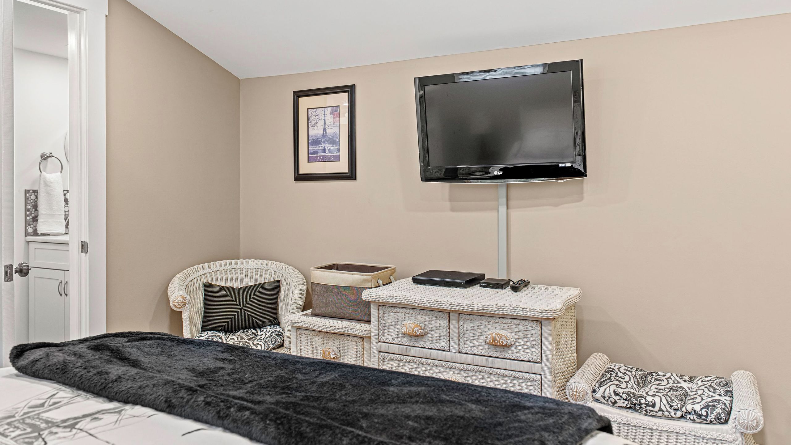 A bedroom with a bed, a wicker chest of drawers, a television mounted on the wall, and a framed picture.