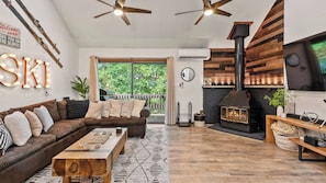 A cozy living room with a sofa, coffee table, and fireplace. There is a large window with a view of greenery outside.