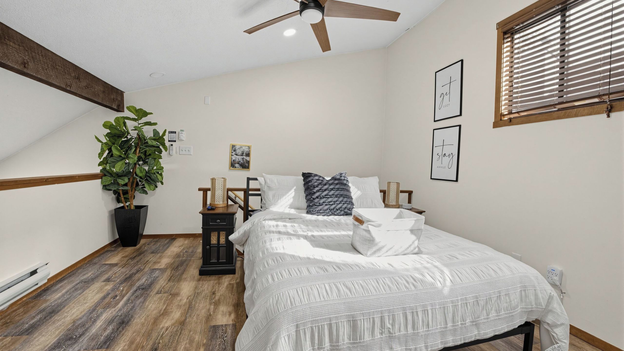 A bedroom with a bed, a nightstand, a potted plant, a ceiling fan, and a window with blinds.