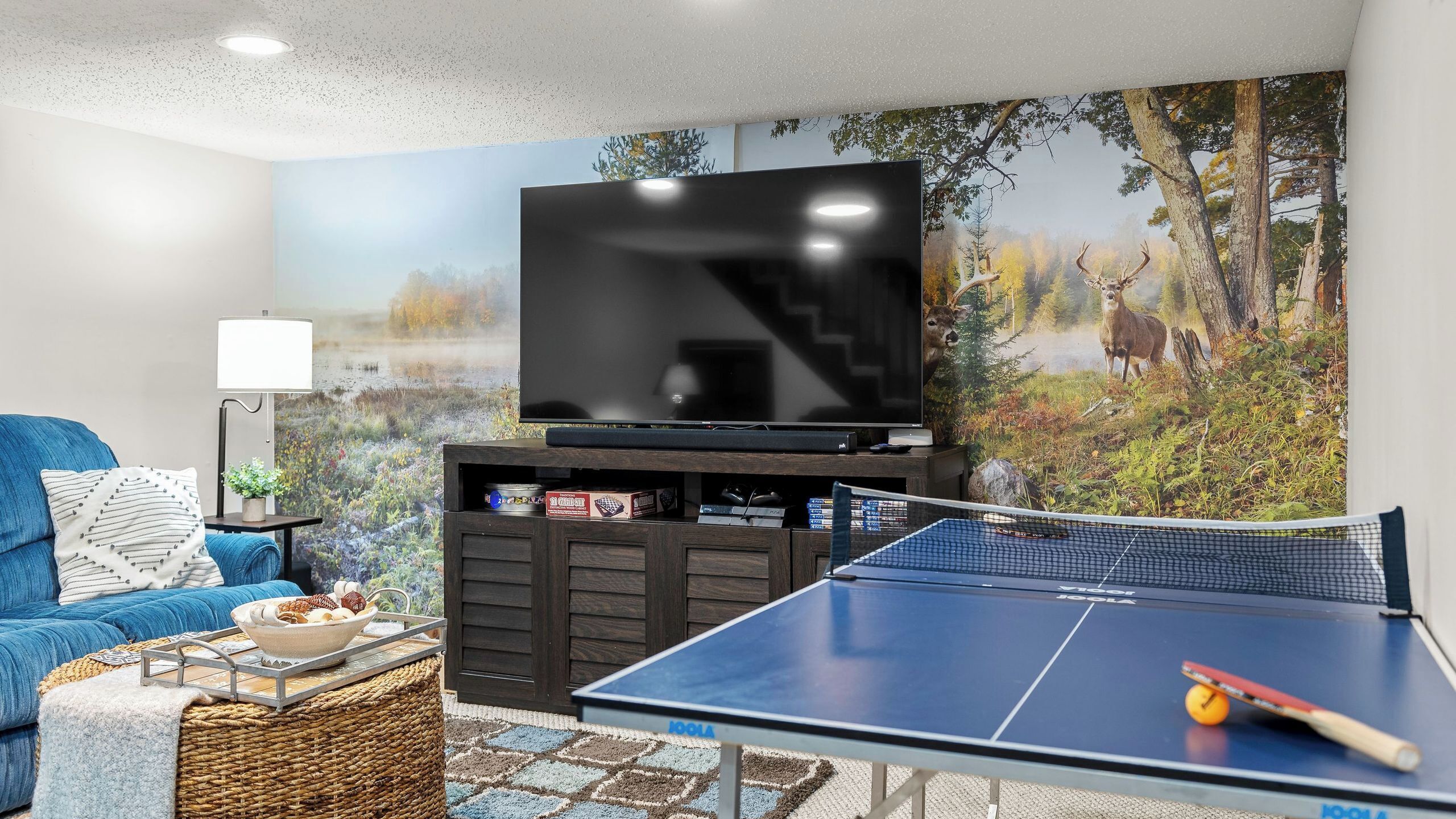 A living room with a TV, a ping pong table, a sofa, and a painting of a forest scene.