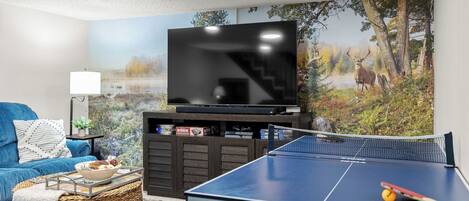 A living room with a TV, a ping pong table, a sofa, and a painting of a forest scene.