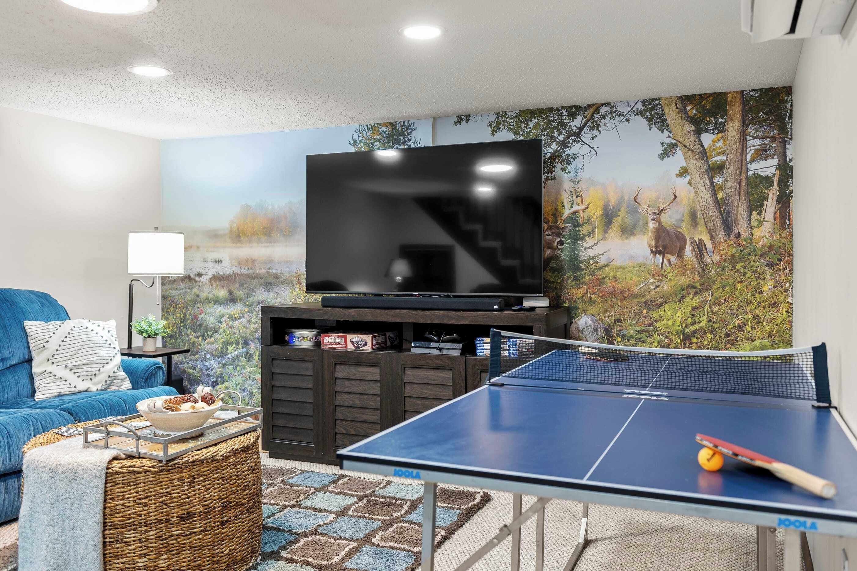 Uma sala de estar com televisão de tela plana, mesa de pingue-pongue, sofá azul e um mural na parede com tema de floresta.