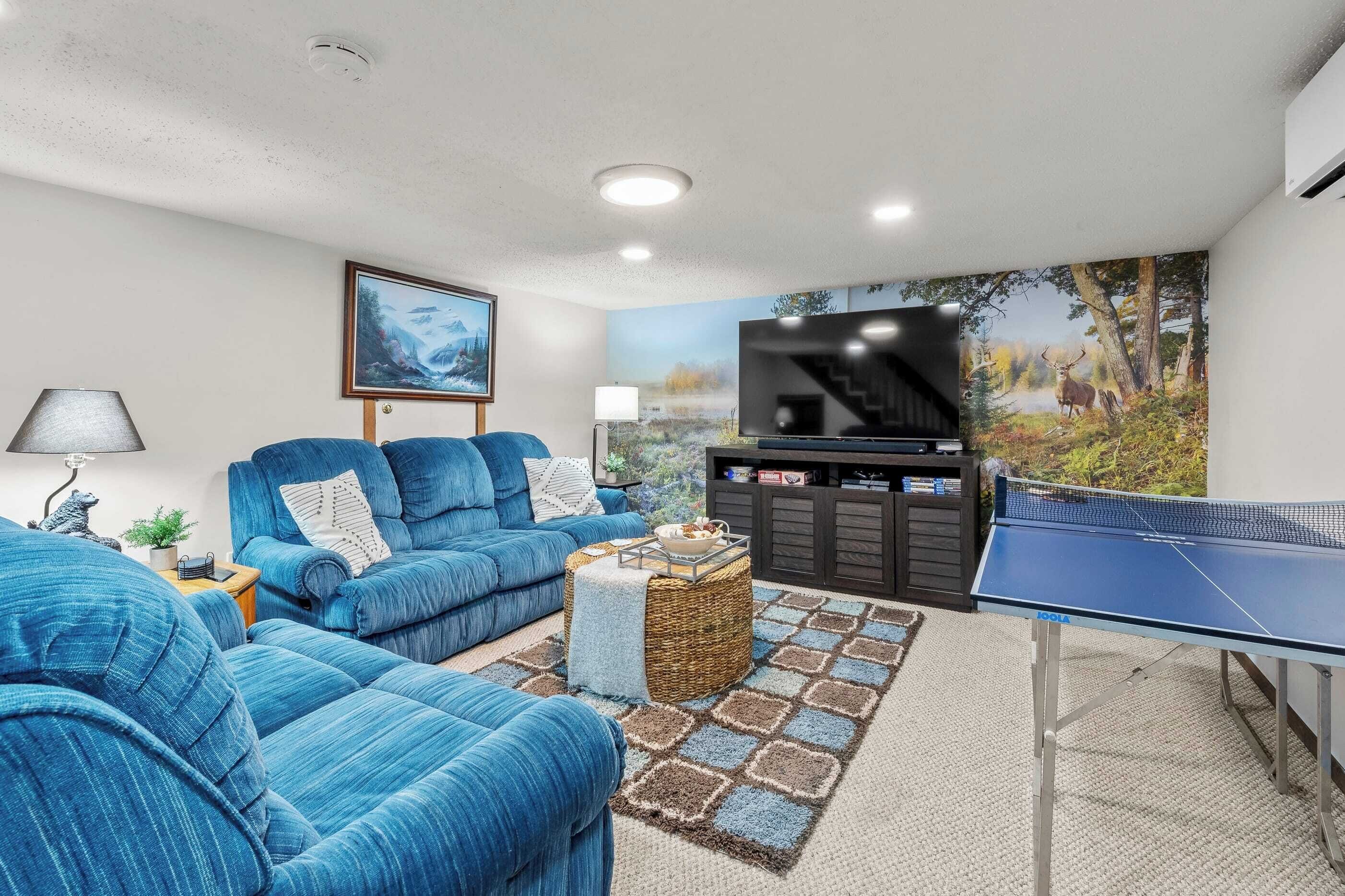 Una sala de estar con un sofá azul, una mesa de ping pong, un televisor y un cuadro de un ciervo en un bosque.