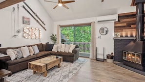 A cozy living room with a sectional sofa, a wooden coffee table, a fireplace, and a large window overlooking greenery.
