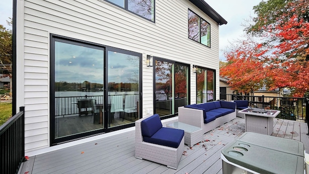 Ein modernes Haus mit großen Schiebeglastüren, einer Terrasse mit Gartenmöbeln und Blick auf einen See.