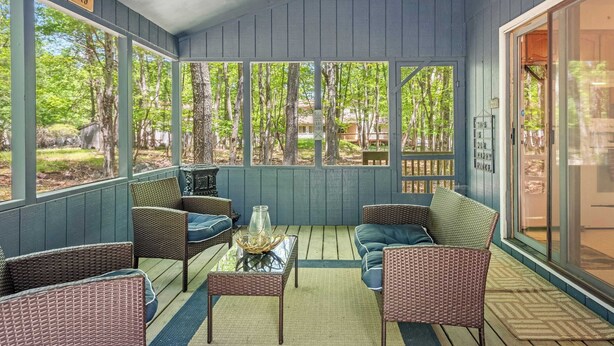 Varanda coberta com móveis de vime, mesa de vidro e vista para árvores e uma casa.