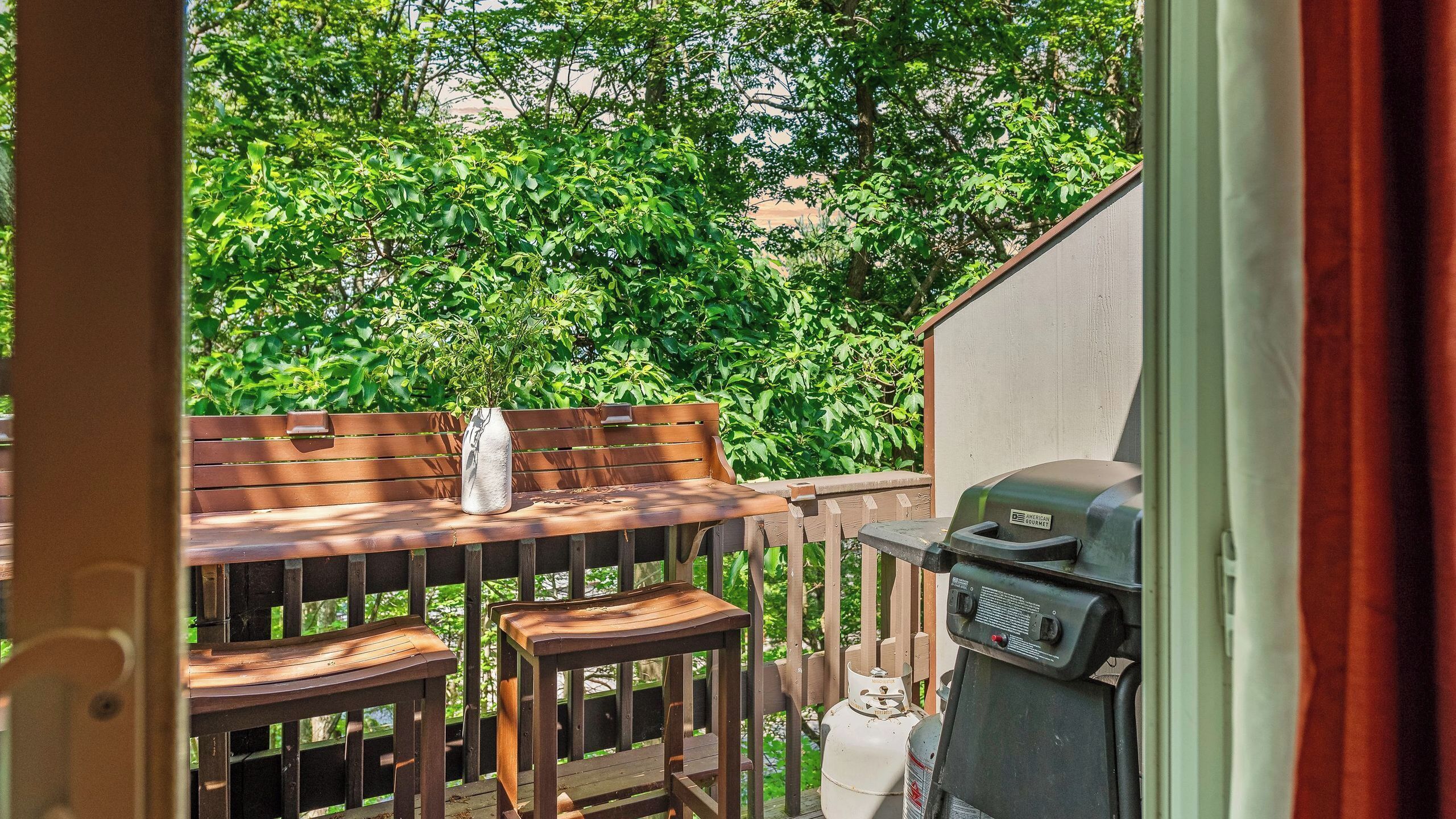 An outdoor patio with a wooden table and chairs, a grill, and lush greenery in the background.