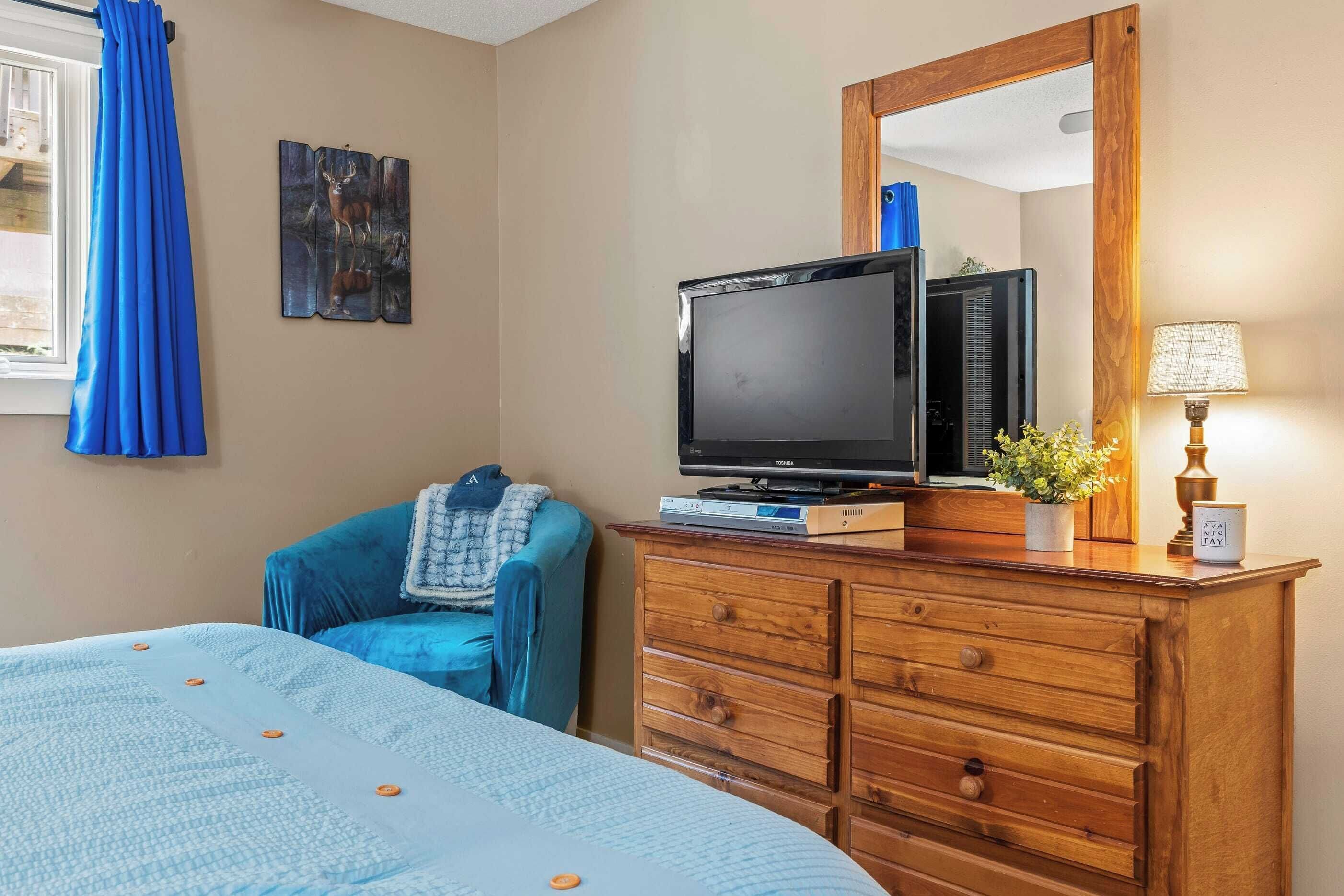 Une chambre d’hôtel avec un lit, une commode en bois, une télévision, un miroir et une fenêtre avec des rideaux bleus.