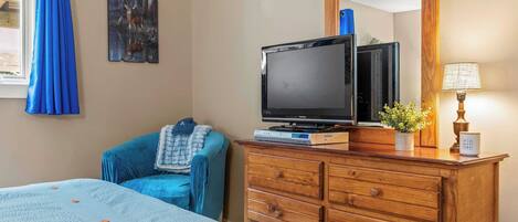 Une chambre d’hôtel avec un lit, une commode en bois, une télévision, un miroir et une fenêtre avec des rideaux bleus.