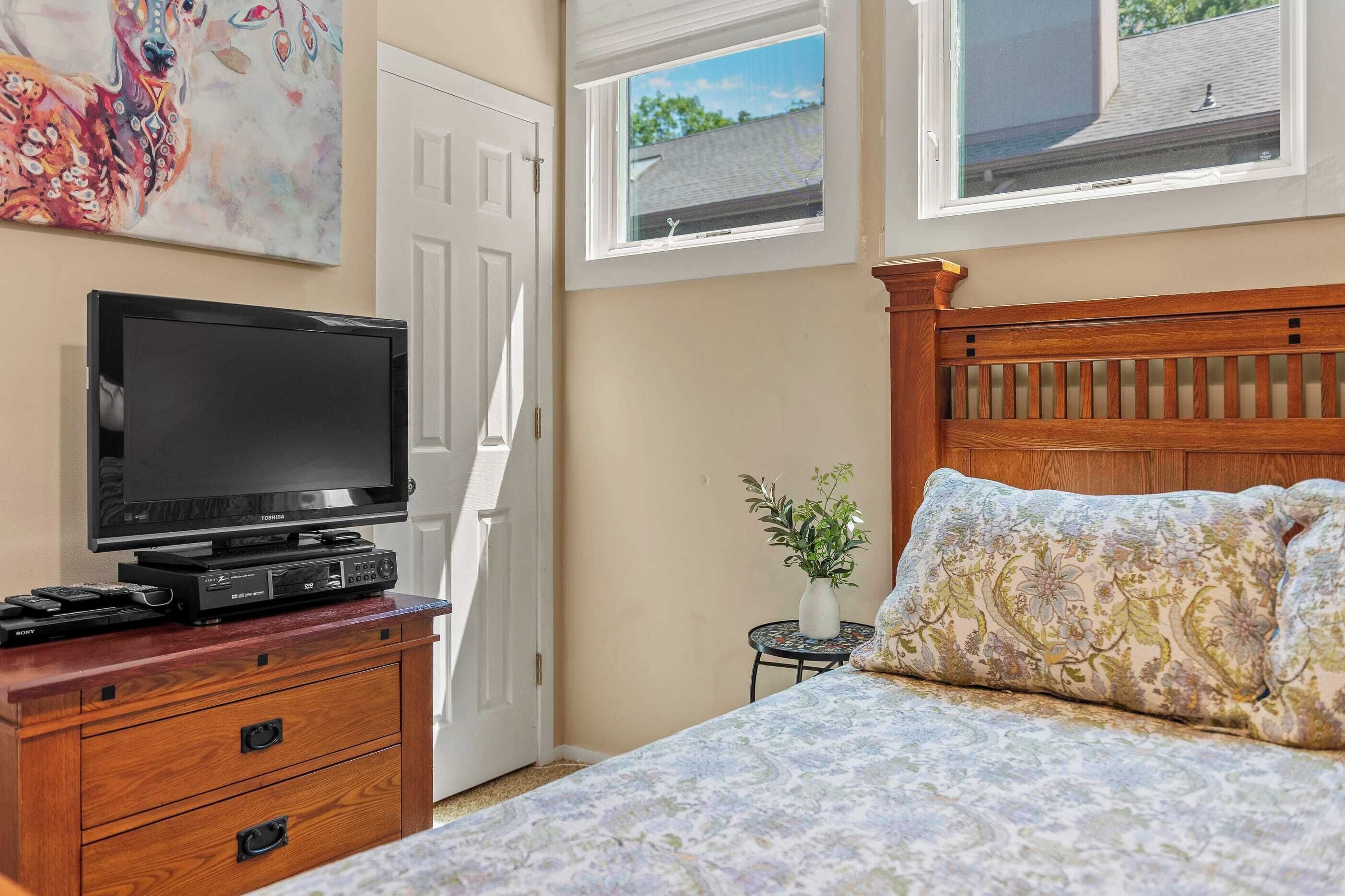 Quarto com cama, televisão sobre uma cômoda de madeira, porta e duas janelas.