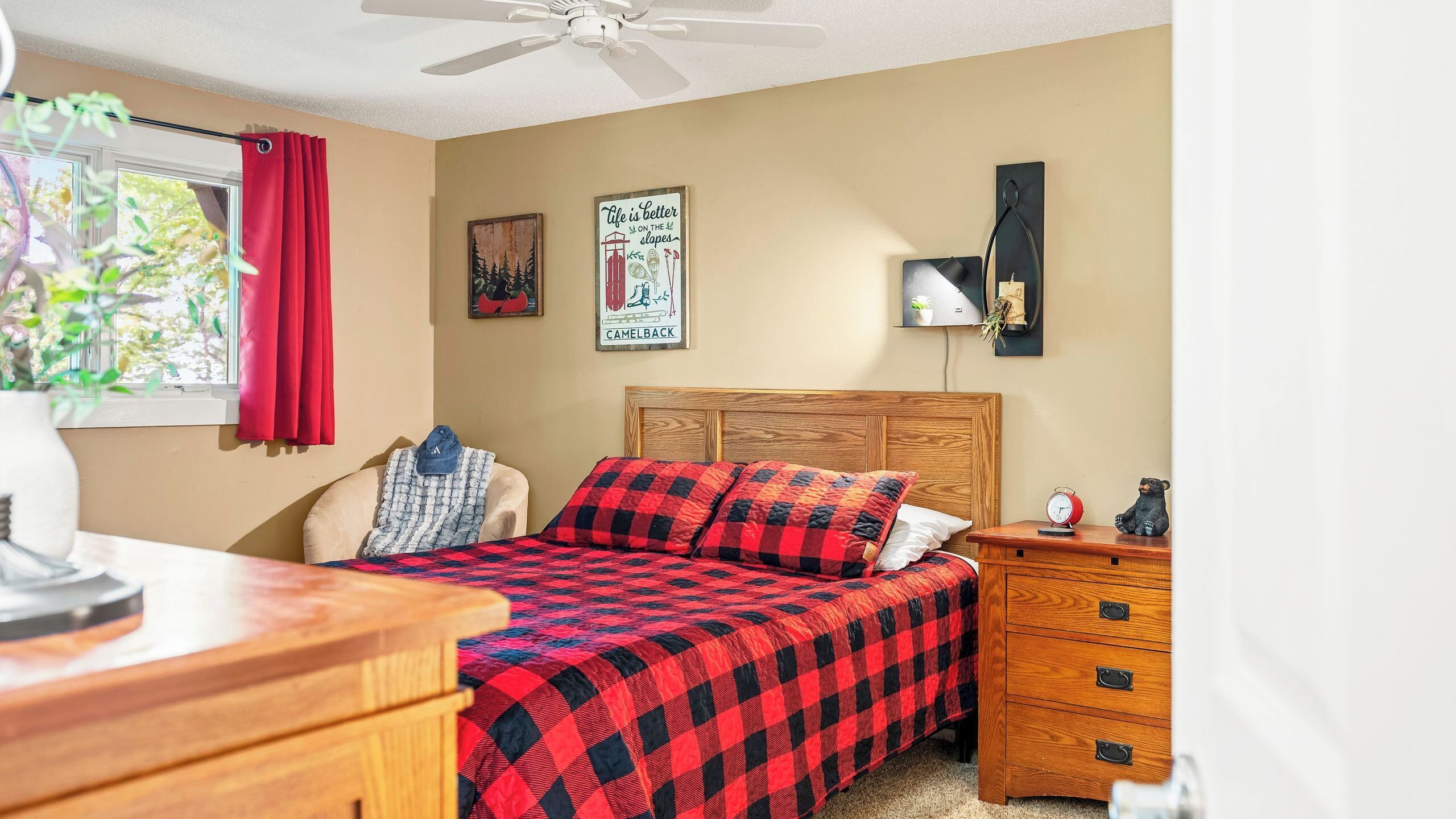 A bedroom with a bed, a nightstand, a dresser, a ceiling fan, a window with red curtains, and a wall with a poster.