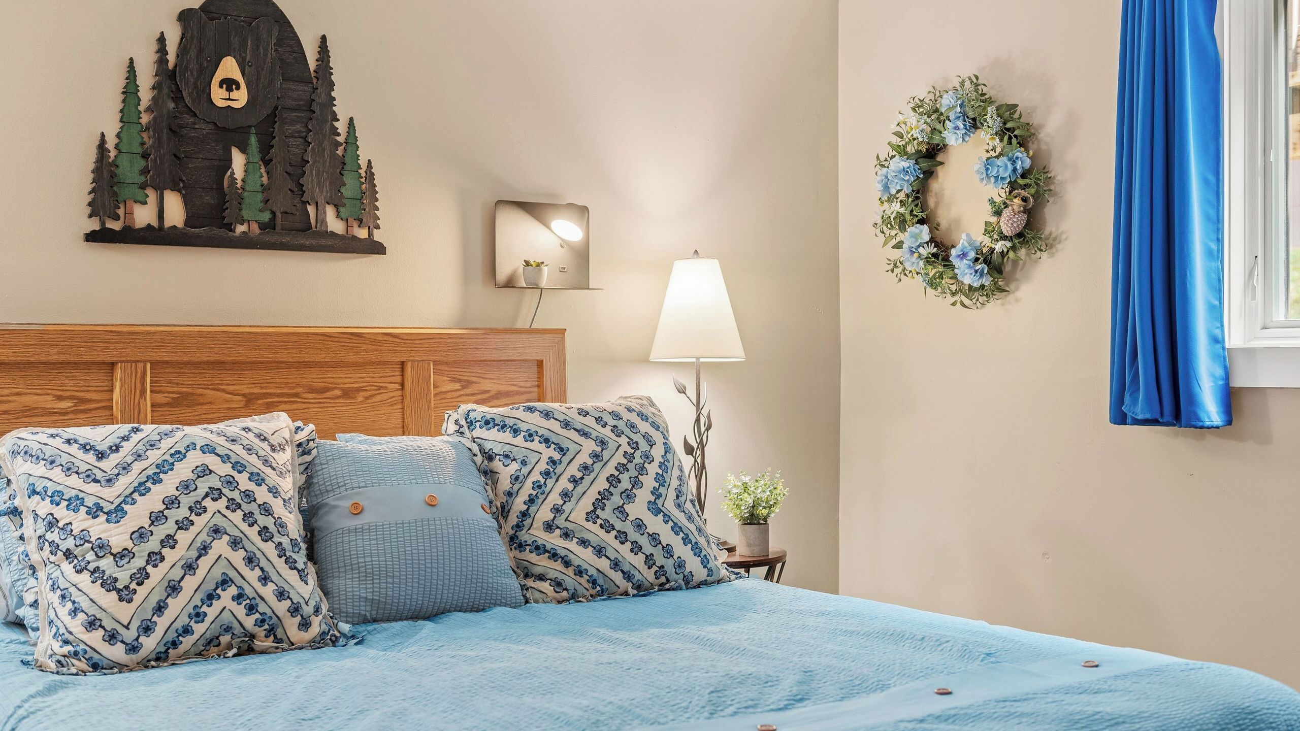 A bed with patterned pillows, a headboard, a wall-mounted lamp, a decorative wreath, and a window with a curtain.