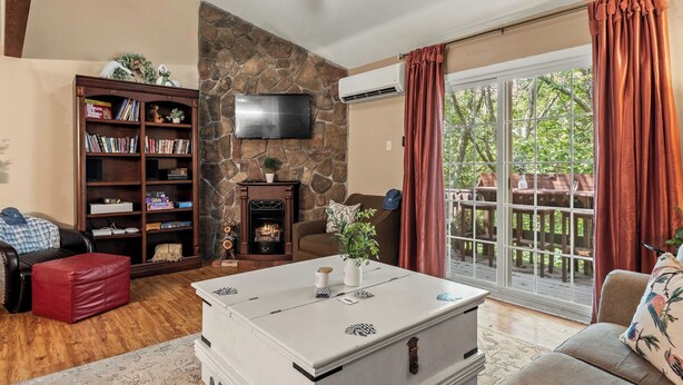 Una sala de estar con chimenea de piedra, una estantería, un televisor de pantalla plana y vistas al exterior a través de amplias ventanas.
