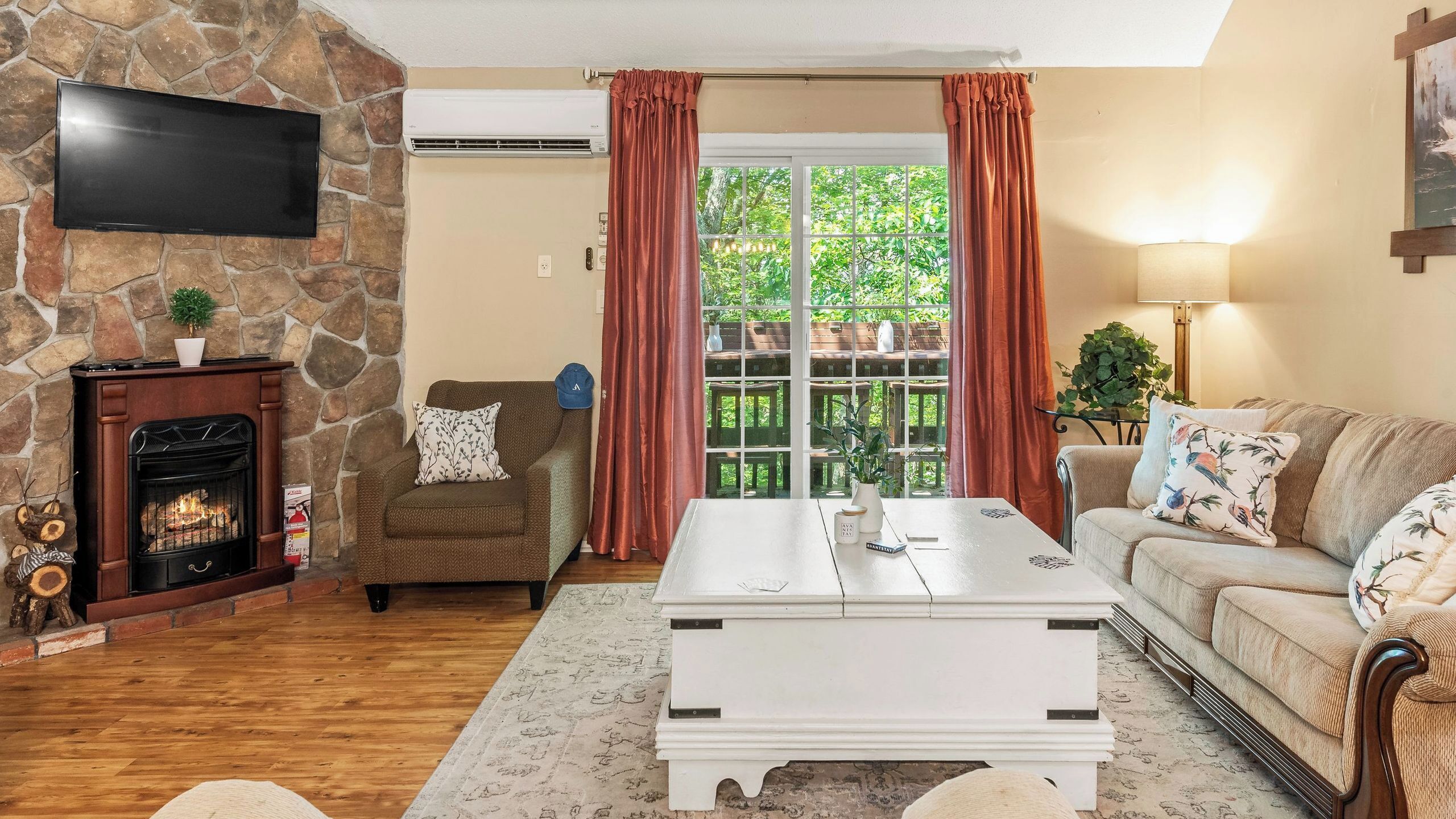 A living room with a stone fireplace, a flat-screen TV, a sofa, a coffee table, and a view through a glass door.