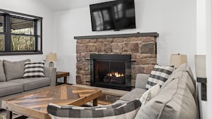 A living room with a stone fireplace, a flat-screen TV mounted above it, a grey sofa, and a wooden coffee table. - The Breck by Avantstay Modern 6bdr Cabin w/ Hot Tub & Game Room (Lake Harmony)