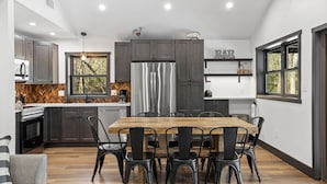 A modern kitchen with dark cabinets, a stainless steel refrigerator, and a wooden dining table with black chairs. - The Breck by Avantstay Modern 6bdr Cabin w/ Hot Tub & Game Room (Lake Harmony)