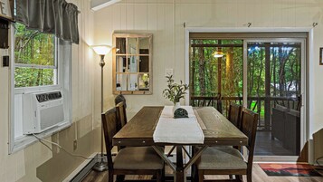 A dining area with a wooden table, chairs, a vase with plants, a lamp, and a view of a forest through a glass door.