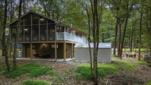 A two-story cabin with a balcony, surrounded by trees and a grassy area.