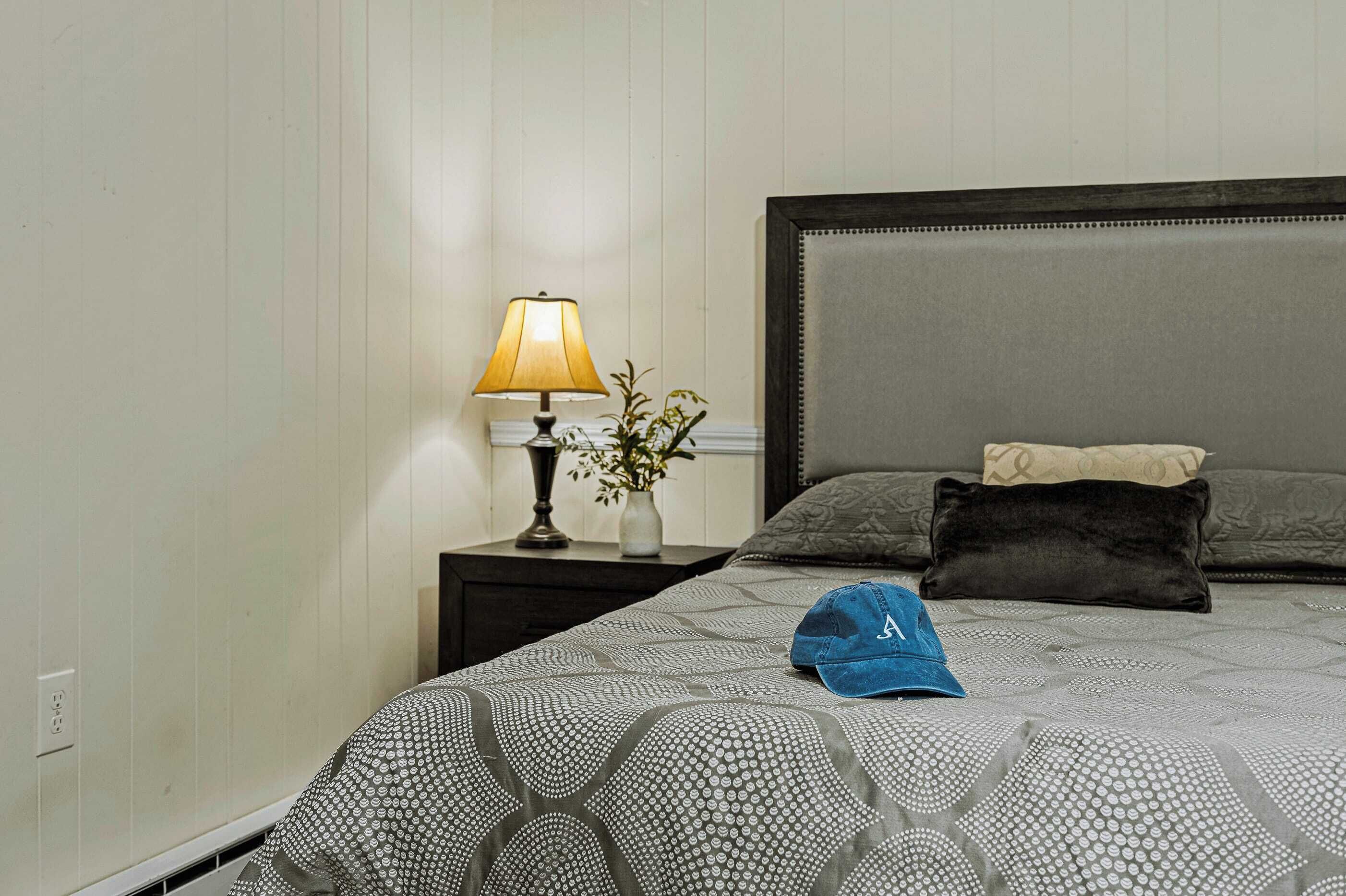 Una cama bien hecha con una colcha estampada, una lámpara de noche, un florero con flores y un gorro azul sobre la cama.