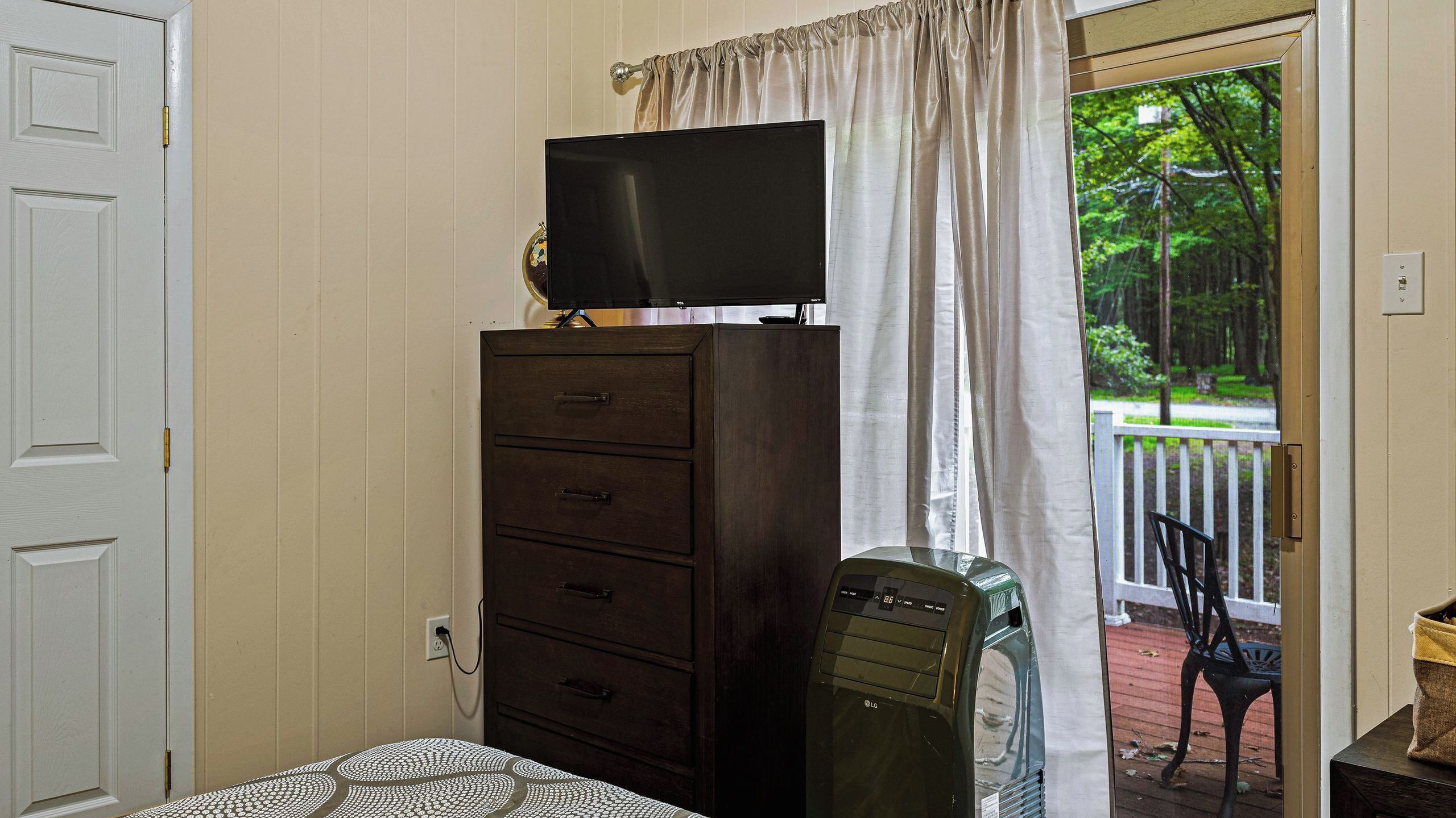 A room with a bed, a dresser, a television on top, a window with curtains, and a view of trees outside.