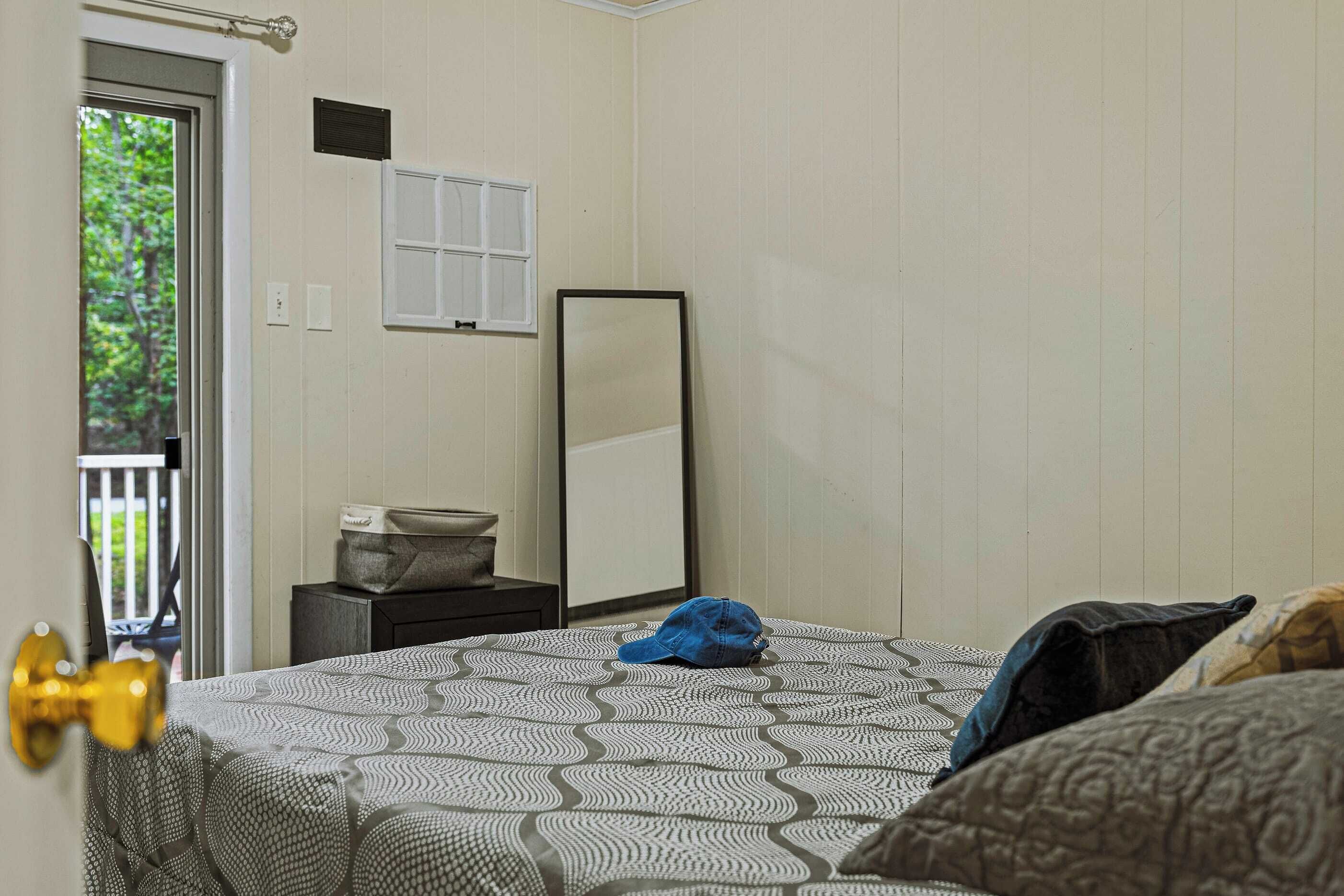 A bedroom with a patterned bedspread, a bedside table with a lamp, a mirror, and a view of greenery through the door.