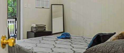 A bedroom with a patterned bedspread, a bedside table with a lamp, a mirror, and a view of greenery through the door.